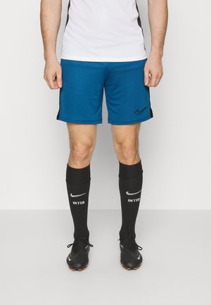 Homme portant un short de sport bleu Nike, des chaussettes de football noires avec le texte "INTER" et des crampons noirs Nike, debout devant un fond blanc uni.