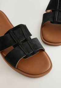 Svarta mockaskinnsandaler med två breda remmar i geometrisk design, en brun vadderad fotbädd och en rund tåsdesign.