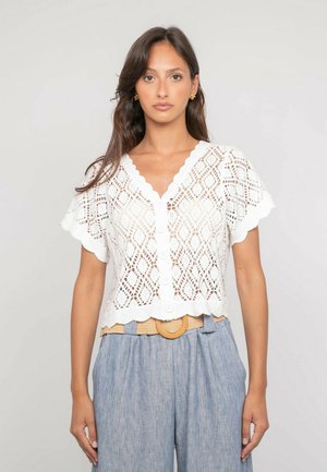 Femme portant un haut blanc en dentelle à manches courtes avec des boutons et des bords en forme de festons, un pantalon taille haute bleu clair et une ceinture beige avec une boucle circulaire.