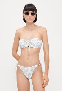 Bikini set s bielym základom a zelenými kvetinovými vzormi, bezramienkový top s šnúrkou a bočnými nohavičkami so zaväzovaním a korálkovými detailmi.