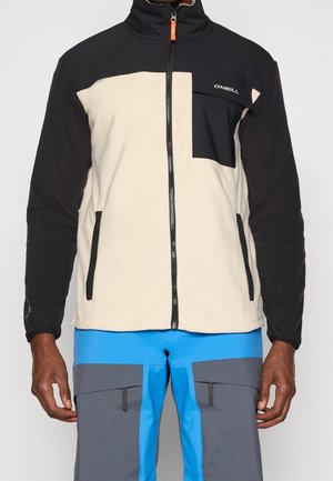 Männlich, trägt eine beige-schwarze Zip-Fleecejacke mit Fronttasche und blaue und graue Outdoor-Hosen mit Reißverschlusstaschen.