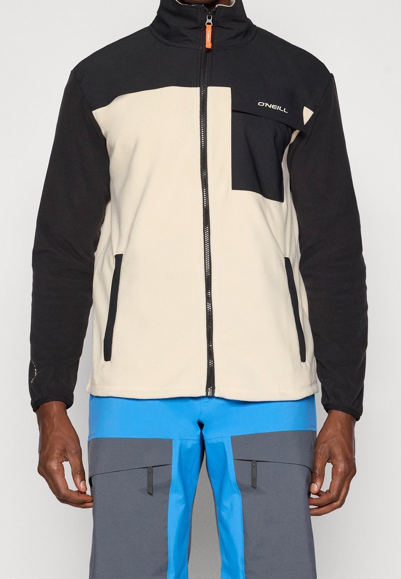 Homme portant une veste polaire zippée beige et noire avec une poche sur le devant et un pantalon d'extérieur bleu et gris avec des poches zippées.