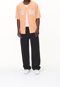 Perzikkleurig overhemd met korte mouwen en knopen, met "PG DR" borduursel, gecombineerd met zwarte wijde broekspijpen en witte sneakers.