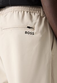 Pantalons de sport beiges avec une taille élastique, dotés d'une poche arrière zippée et d'un logo noir "BOSS" imprimé près de la poche.
