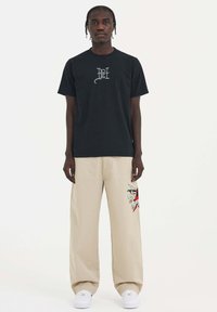 Czarna bawełniana koszulka z graficznym nadrukiem na klatce piersiowej, zestawiona z beżowymi spodniami o szerokich nogawkach z haftowanym wzorem i białymi trampkami.
