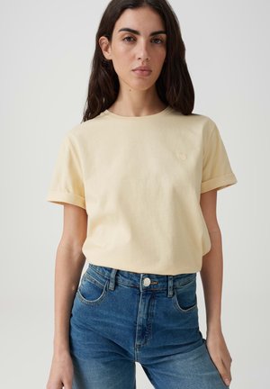 Donna con i capelli lunghi e scuri che indossa una camicia giallo pallido a maniche corte infilata in jeans blu a vita alta, in piedi davanti a uno sfondo chiaro e semplice.