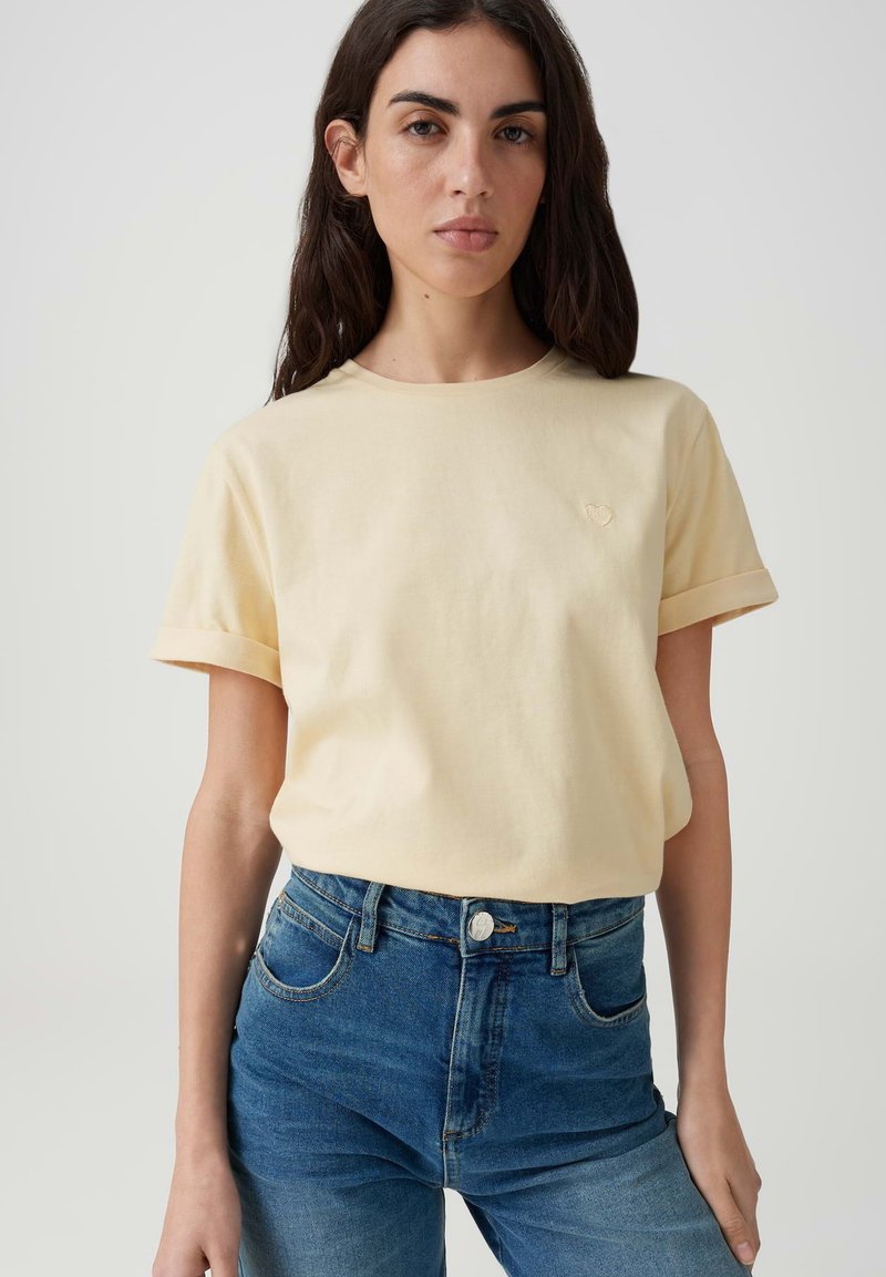 Donna con i capelli lunghi e scuri che indossa una camicia giallo pallido a maniche corte infilata in jeans blu a vita alta, in piedi davanti a uno sfondo chiaro e semplice.