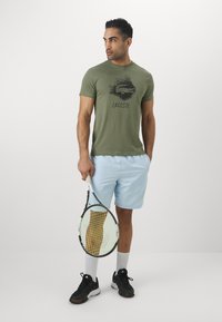 Green cotton t-shirt with black Lacoste logo, paired with light blue shorts and black athletic shoes. Holding a tennis racket.