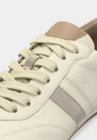 Δερμάτινο αθλητικό παπούτσι σε εκρού απόχρωση με μπεζ λεπτομέρεια στη φτέρνα, με υφή στην επιφάνεια, επίπεδα κορδόνια και ανοιχτογκρι ρίγα στο πλάι.