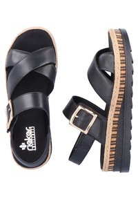 Des sandales en cuir noir présentent des lanières croisées, une boucle de cheville ajustable, et une semelle intermédiaire en jute tissé avec une semelle extérieure texturée noire.