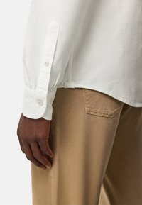 Långärmad vit skjorta med knäppningar vid ärmsluten, parat med ljusbruna byxor. Skjortan har en avslappnad passform och texturerad tyg.