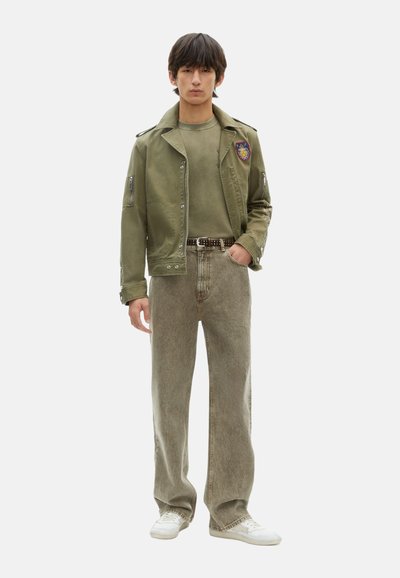 Olijfgroene jas met patches, bijpassende olijfgroene shirt en licht beige wijdvallende jeans; afgewerkt met witte sneakers. Gevarieerde stoffen doorheen.