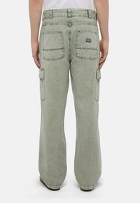 Osoba v volných, vybledlých zelených cargo kalhotách s velkými zadními kapsami a nášivkou loga na pravé kapse, stojící na bílém pozadí.