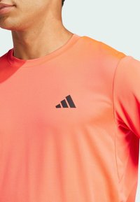 Camiseta deportiva naranja hecha de tela transpirable, con un escote redondo y un logo negro en el pecho. Textura suave con un diseño ajustado.