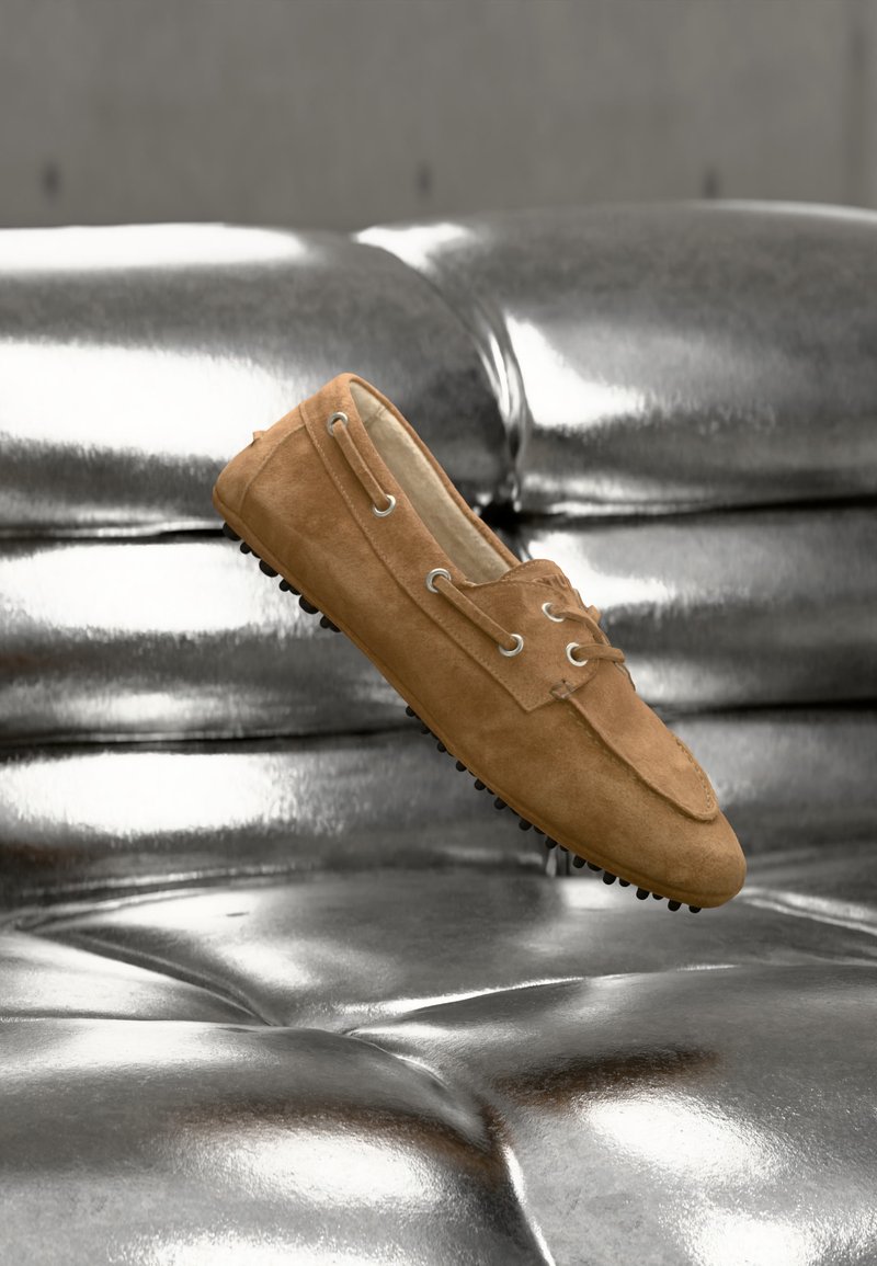 Mocassin en daim marron avec semelle en caoutchouc noire reposant sur une surface métallique argentée brillante et rembourrée.