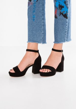 Mustat mokkanahkaiset korolliset sandaalit, joissa leveä nauha varpaiden päällä ja nilkkahihna. Varustettu paksulla lohkokorkokengällä ja avoimella varvasosalla.