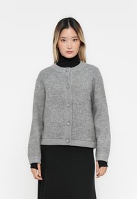 Šedý pletený cardigan s kulatým výstřihem, vpředu na knoflíky a žebrovanými manžetami. Měkká textura, zkrácený design v kombinaci s černým topem.