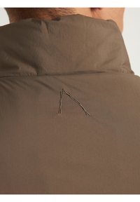 Jakna khaki boje s visokim ovratnikom. Značajna karakteristika: unikatni dizajn u obliku 'A' izvezen na leđima, sa teksturiranim površinom tkanine.