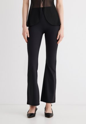 Naine, kes kannab musta kitsast laiade säärega pükse, musta läbipaistvat pluusi ja musti teravate nina ning rihmaga kingi, seisab valgel põrandal ühevärvilise tausta ees.