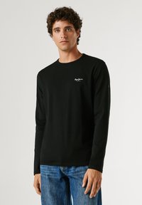 Mladi moški z kodrastimi lasmi, oblečen v črno majico z dolgimi rokavi z majhnim belim logotipom in modre kavbojke, stoji pred enobarvnim ozadjem.