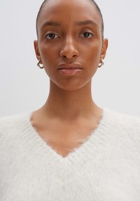 Weißer, flauschiger V-Ausschnitt-Pullover mit weicher Textur. Das Model trägt kleine silberne Creolen, vor einem hellen Hintergrund, der das Kleidungsstück betont.