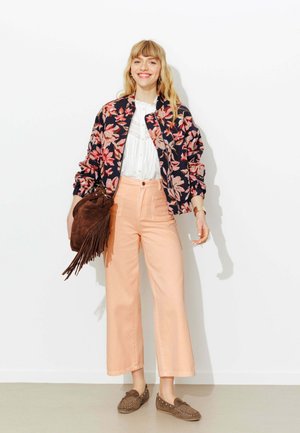 Femme souriante portant une veste bomber fleurie, un chemisier blanc, un pantalon ample pêche, des mocassins marron, tenant un sac en daim marron à franges contre un mur blanc.