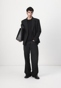 Schwarze Blazer über einem schwarzen T-Shirt, kombiniert mit lockeren schwarzen Jeans und glänzenden schwarzen Schuhen. Die Person hält eine elegante schwarze Umhängetasche.