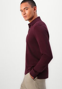 Burgundy long-sleeve polo shirt with a collared neck, buttons down the front, and ribbed cuffs. Made of textured fabric.