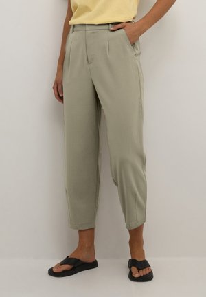 Femme portant un pantalon fuselé taille haute vert clair, un haut jaune et des sandales noires à bout ouvert, debout avec une main dans la poche.