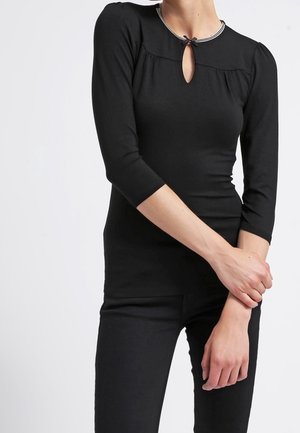 Femme portant un haut noir ajusté avec manches trois-quarts, encolure en forme de trou clé, petit détail nœud et pantalon noir, debout devant un fond blanc.