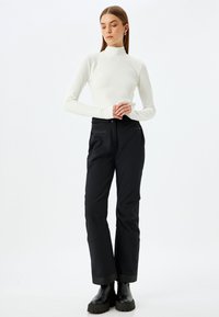 Witte langwerpige turtleneck top gecombineerd met zwarte broek. De broek heeft een zachte textuur, is lang en heeft zichtbare voorzakken. Zwarte laarzen.