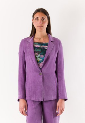 Donna che indossa una giacca e pantaloni da completo viola con una maglia floreale multicolore, in piedi davanti a uno sfondo bianco semplice.