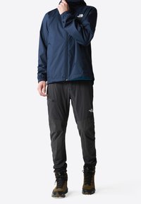 Chaqueta impermeable azul marino con puños elásticos, combinada con pantalones negros con bolsillos y botas de senderismo marrones con cordones negros.