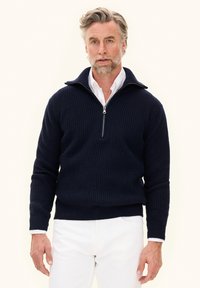 Uomo con capelli e barba grigi che indossa un maglione navy a mezza zip sopra una camicia bianca e pantaloni bianchi, in piedi su uno sfondo chiaro.