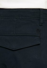 Dunkelblaue Jeanshose mit einer hinteren aufgesetzten Tasche mit dreieckigem Klappdeckel und gesticktem Logo-Detail in der Nähe des Bundes.