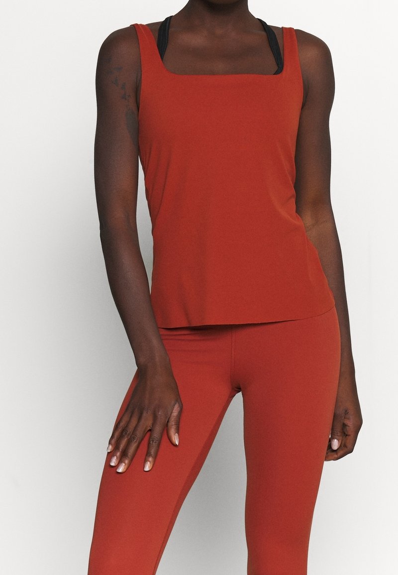 Femme portant un haut sans manches couleur rouille et un legging assorti, avec des bretelles visibles de brassière de sport noire, debout devant un fond clair uni.