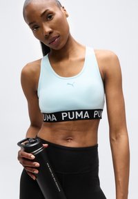 Αθλητική γυναίκα με ανοιχτομπλε αθλητικό σουτιέν Puma και μαύρα κολάν, κρατώντας μια μαύρη φιάλη νερού μπροστά σε ουδέτερο φόντο.