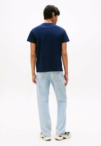 Joven de pie de espaldas, vestido con una camiseta azul marino, jeans claros y zapatillas blancas sobre un fondo blanco.