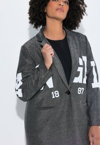 Frau mit lockigem Haar, die ein texturiertes graues Blazer mit weißem, fettem Text und Zahlen über einem schwarzen Top trägt, posiert vor einem weißen Hintergrund.