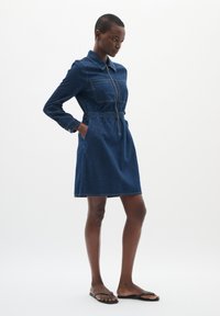 Denim jurk met een kraaghalssluiting, lange mouwen, ritssluiting aan de voorkant, elastische tailleband en twee zijzakken. Gedragen met platte sandalen.
