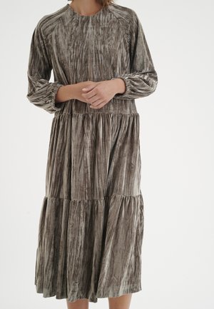 Mujer vestida con un vestido midi de manga larga y volantes, con una suave textura de terciopelo aplastado en tonos gris y marrón apagados, con las manos unidas en la cintura.
