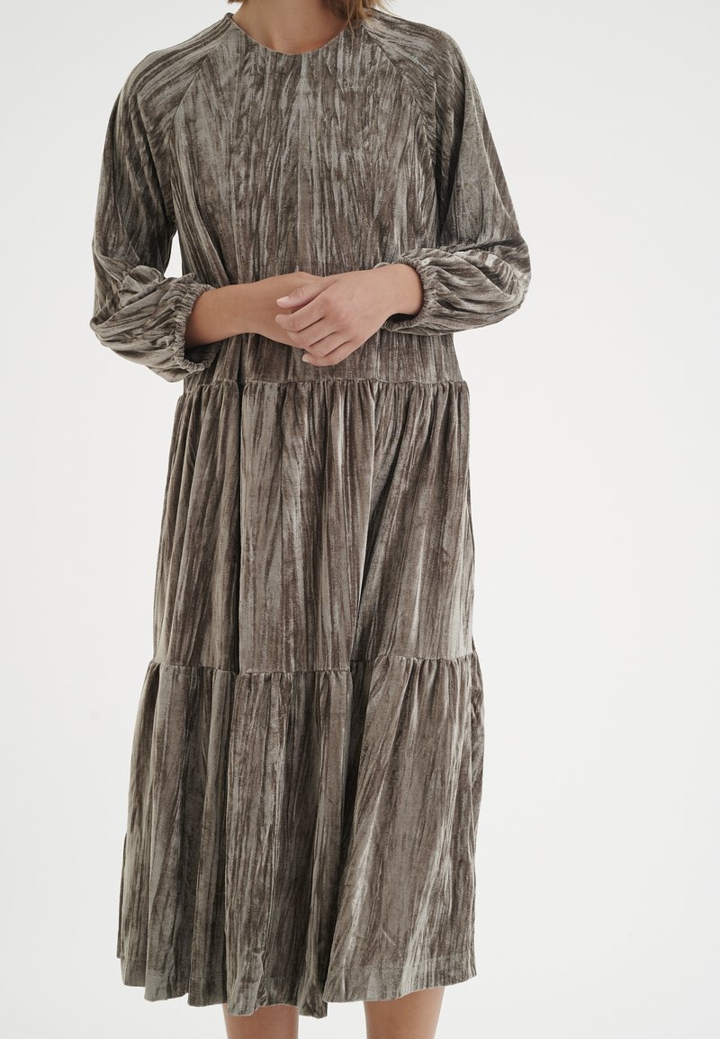 Kvinna i långärmad, lager-på-lager midi-klänning med mjuk, krossad sammetstextur i dämpade gråa och bruna toner, händerna sammanlagda vid midjan.