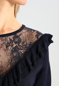 Marinepullover mit Spitze überzogen, die florale Muster zeigt. Rüschendetails entlang der Schulternaht sorgen für Textur und Designinteresse.