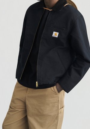 Person trägt eine schwarze Zip-up-Jacke mit einem Logopatch auf der Brust und beige Hose, die Hände lässig in den Taschen vor einfachem Hintergrund.