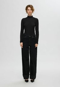 Juodas ilgomis rankovėmis viršus su tekstūruotu raštu, derinamas su aukštu liemeniu juodomis plačiomis džinsinėmis kelnėmis su sukta apatine dalimi ir smailiais juodais batais.