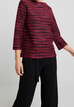 Kobieta w czerwono-czarnym teksturowanym pasiastym topie z długim rękawem, w połączeniu z luźnymi czarnymi spodniami, na jednolitym jasnoszarym tle.