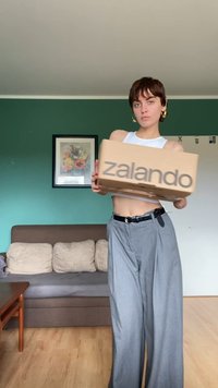 Kartonska škatla z napisom "zalando" v rjavi barvi na sprednji strani. Model nosi bel kratek top in ohlapne sive hlače, stoji v dnevni sobi.