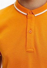 Oranžové polo tričko se strukturovanou textilií, bílým pruhovaným límečkem, dvoubotónovým zapínáním a jednou náprsní kapsou.