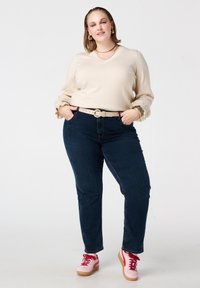 Beige trui met V-hals en gerimpelde manchetten, donkerblauwe spijkerbroek, lichte riem en roze sneakers met rode veters, geplaatst tegen een eenvoudige achtergrond.