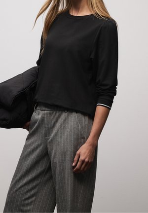 Kobieta ubrana w czarną koszulę z długim rękawem z białym obramowaniem na mankiecie oraz szare spodnie w prążki, trzymająca czarną pikowaną kurtkę na jednym ramieniu.